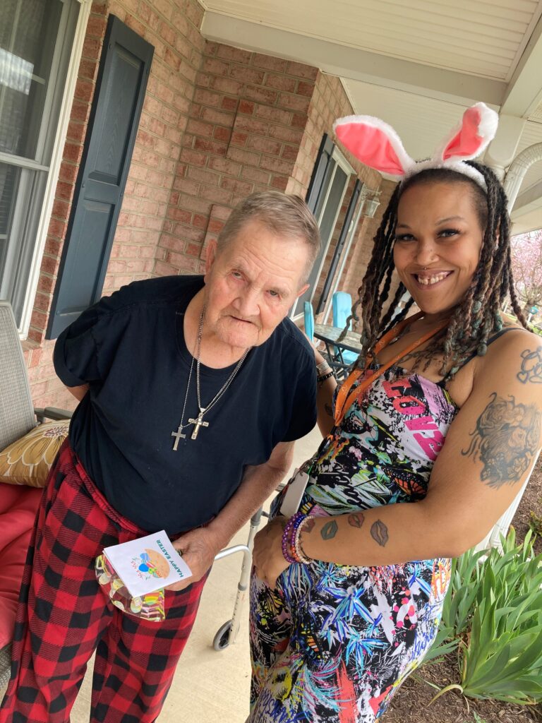 A man on a porch holds some Easter goodies as a woman wearing a bunny ear headband smiles with her arm around him.
