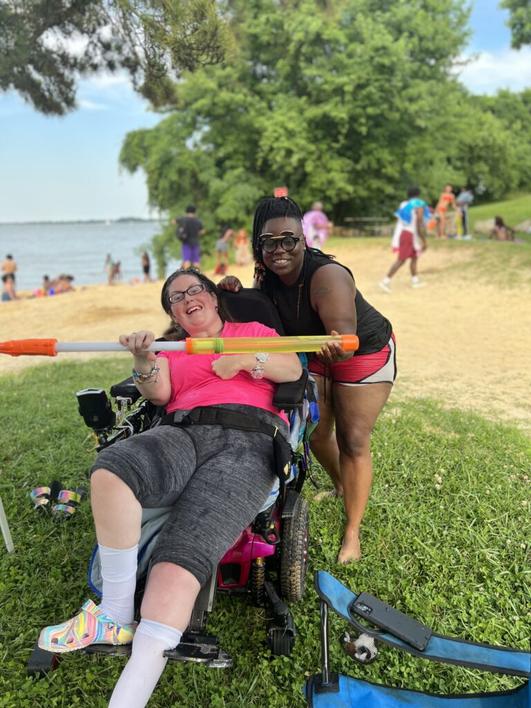 A woman in a power chair and a woman standing behind her smile as the hold up a water gun. They are at a beach.