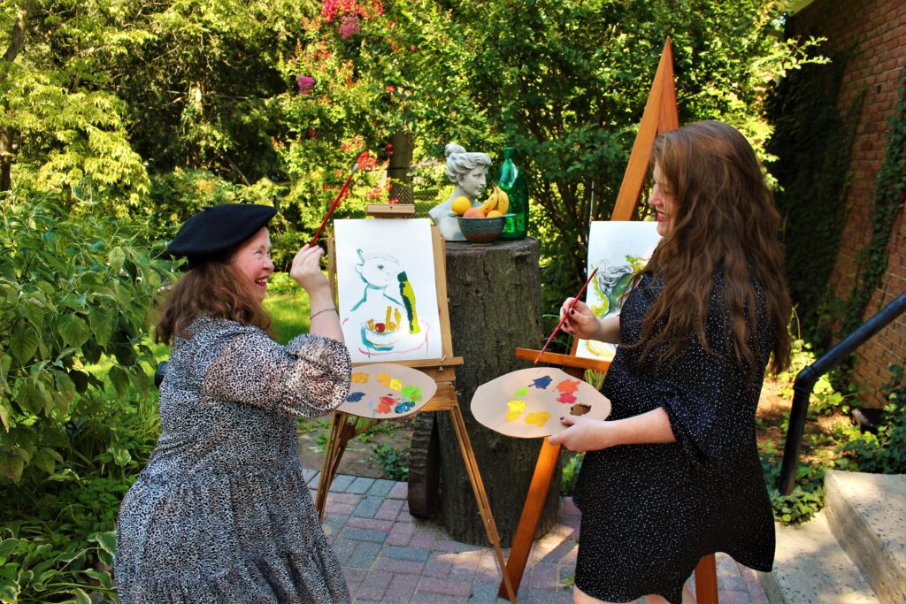 Two women smile at each other in a yard holding their palettes and paint brushes in front of their canvasses.