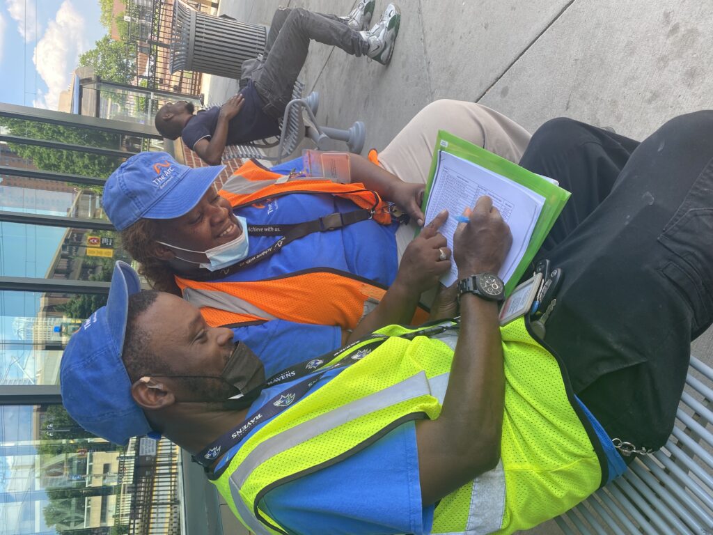 A man and woman smiling and wearing high visibility safety vests sit and look down at a document they are reviewing while sitting on a bench outside.